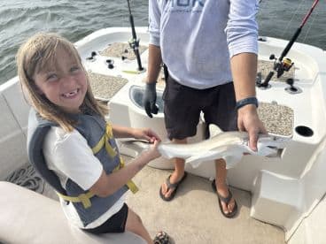 A young girl in a life jacket joyfully holds a caught fish while on a fishing boat, showcasing a fun family fishing experience.