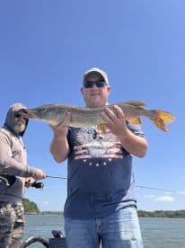 An angler proudly displays a large Northern Pike while fishing on a sunny day, with another fisherman in the background.