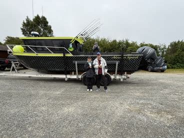 A couple stands beside the 'Alaska Spirit' boat, showcasing its unique design and features in a scenic outdoor setting.