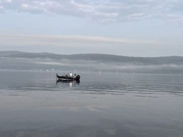 A fishing boat is seen gliding across a calm lake, surrounded by misty hills, creating a serene fishing environment.
