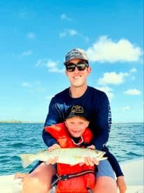 A young boy and an adult proudly displaying a Speckled Trout while enjoying a fishing trip on a sunny day.