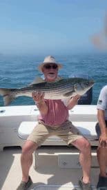 An angler proudly displaying a large Striped Bass while fishing on a boat, showcasing a successful day on the water.