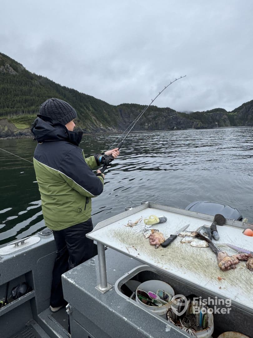 A young angler is actively reeling in a catch while fishing from a boat in a scenic coastal environment, with rocky cliffs and lush greenery in the background.