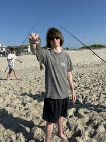 A young angler proudly displaying a caught fish on the beach, with fishing rods in the background, showcasing a fun day of fishing.