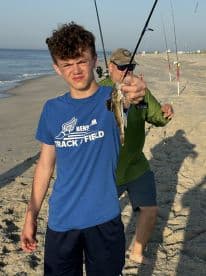 A young angler proudly displaying a caught fish on the beach, with fishing rods set up in the background, capturing a lively fishing experience.