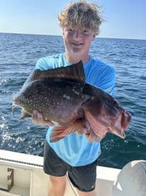 A young angler proudly displaying a large Grouper while fishing offshore, showcasing a sunny day on the water.