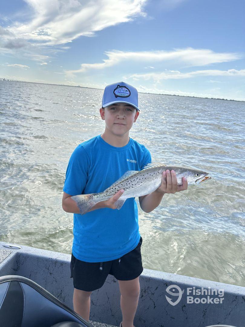 Young angler proudly holding a Speckled Trout while fishing on a boat in a coastal waterway.