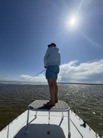 A lone angler standing on a casting platform of a boat, casting into the water under a bright sky, showcasing a serene fishing environment.