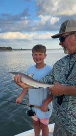 A young angler proudly holding a small shark while fishing with an adult on a calm waterway.