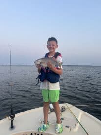 A young angler proudly displaying a caught fish while standing on a boat in calm waters, showcasing a fun fishing experience.