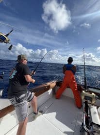 Two anglers actively fishing on a sportfishing boat, with one reeling in a line while the other prepares for a catch in a vibrant ocean setting.