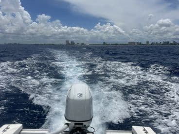 A scenic view from the back of a boat, showcasing the wake and skyline of a coastal city under a partly cloudy sky.