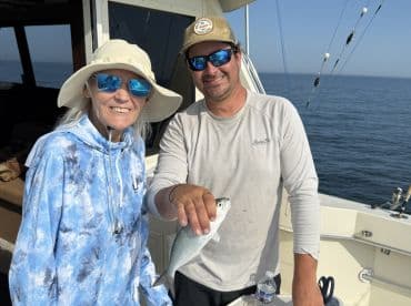 Two anglers smiling on a boat, showcasing a small fish caught during a day of fishing on the open water.