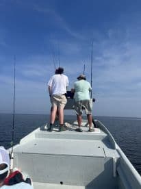 Two anglers engaged in fishing on a boat, with multiple fishing rods set up against a clear blue sky.