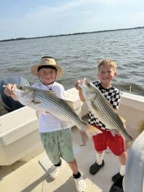 Two young anglers proudly displaying their catch of striped bass while enjoying a sunny day on the water.