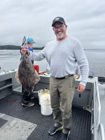 An angler proudly displaying a large Halibut on a fishing boat in a scenic coastal environment.