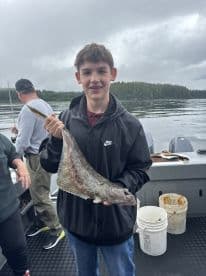 A young angler proudly displaying a Halibut while fishing on a boat in a scenic coastal environment.