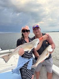 Two anglers proudly displaying their catch of Redfish while on a fishing trip, with a scenic backdrop of the water and cloudy sky.