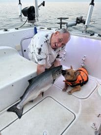 An excited angler poses with a large Salmon while a French Bulldog in a life jacket looks on, showcasing a fun day of fishing on the water.