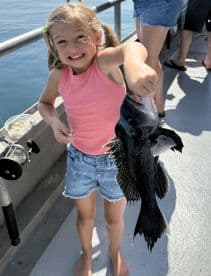 A young girl joyfully displaying her catch, a Black Sea Bass, while fishing on a sunny day.