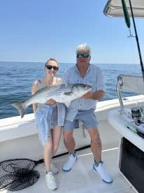 Two anglers proudly displaying a large Striped Bass while fishing on a boat in clear waters.