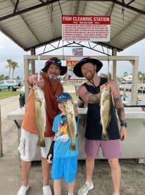 A family proudly displaying their catch of three Speckled Trout at a fish cleaning station after a successful day of fishing.