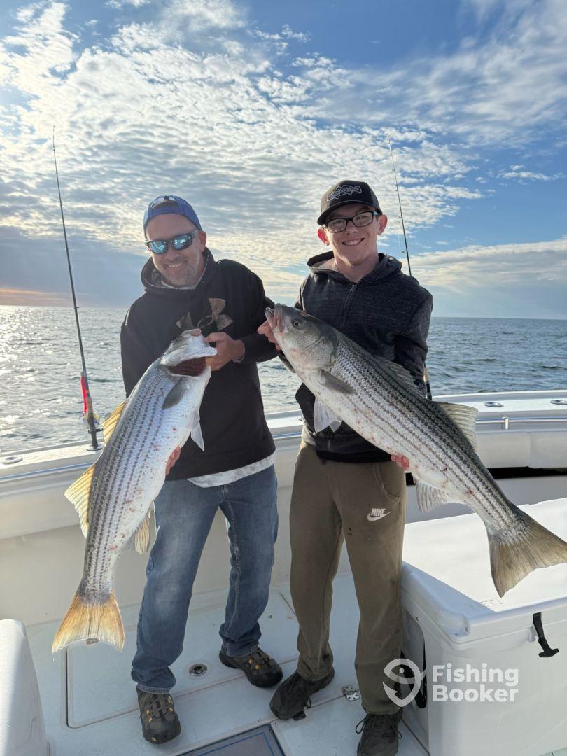 Two anglers proudly displaying their large striped bass while fishing on a boat, enjoying a successful day on the water.