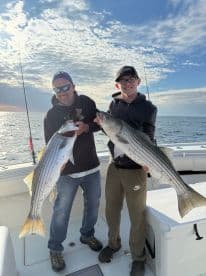 Two anglers proudly displaying their large striped bass while fishing on a boat, enjoying a successful day on the water.