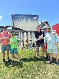 A group of anglers, including a young boy, proudly displaying their catch of the day, featuring several fish, in front of a promotional sign in Rockport, Texas.