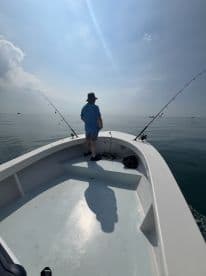 A lone angler stands on the deck of a fishing boat, casting lines into calm waters under a bright sky.