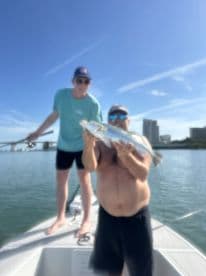 Two anglers on a boat showcasing a large Speckled Trout against a scenic waterfront backdrop.