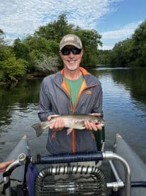 A smiling angler proudly displaying a Rainbow Trout while fishing on a serene river, surrounded by lush greenery.