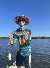 Angler proudly displaying a Speckled Trout while fishing from a boat in a calm waterway, wearing a patriotic hat and fishing gear.
