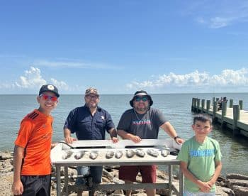 A group of three individuals, including two young boys, proudly displaying their catch of fish on a board at the dock, with a scenic waterfront backdrop.