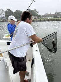 Two anglers actively fishing from a boat, with one using a net to catch a fish in a calm water setting.
