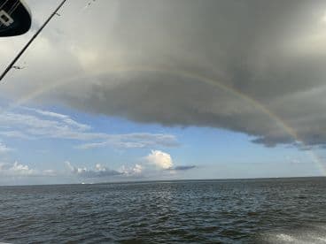 A scenic view of a rainbow arching over the ocean, with clouds and a hint of a boat in the foreground.