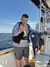 A young angler proudly displaying a speckled trout while fishing on a boat, showcasing a sunny day on the water.