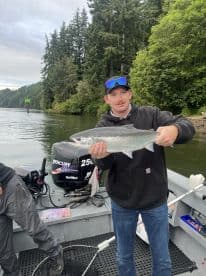 Angler proudly displaying a Silver Salmon while fishing in a scenic river surrounded by lush greenery.