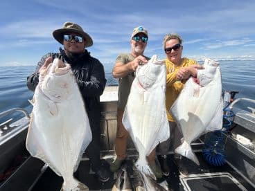 Three anglers proudly displaying their catch of large Halibut while aboard a fishing boat in calm waters.