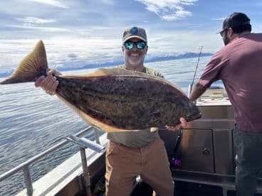 An angler proudly displaying a large Halibut while fishing in open waters, with scenic mountains in the background.