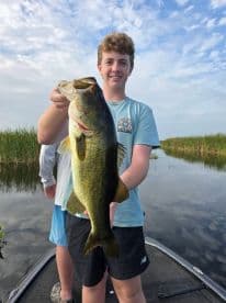 A young angler proudly displaying a large Largemouth Bass while fishing on a serene lake, surrounded by lush vegetation.