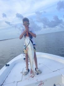 A young angler proudly displaying a large Redfish while standing on a boat in calm waters, showcasing a successful fishing trip.