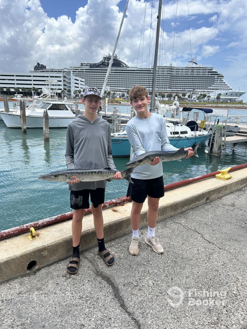 Two young anglers proudly displaying their Barracuda catch at the dock, with a cruise ship in the background.