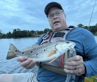 An angler proudly displaying a large Speckled Trout while kayaking on a calm lake, showcasing a successful fishing outing.