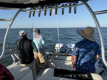 Group of anglers actively fishing on a boat, with one person reeling in a catch while others observe the action on a sunny day.