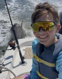 A young angler smiling for a selfie on a boat, with a dolphin visible in the background, capturing a fun moment on the water.