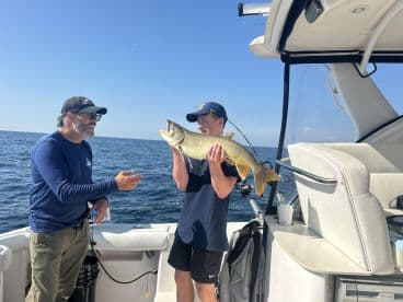A young angler proudly displays a large Lake Trout while fishing on a boat, with a clear blue sky and calm waters in the background.