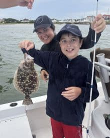 A young boy proudly displaying a Flounder while fishing with an adult on a boat, showcasing a fun family fishing experience.