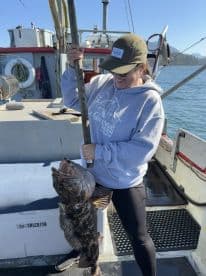 An angler proudly displaying a large Lingcod on a fishing boat, showcasing a successful catch during a Pacific Northwest fishing trip.