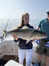 A young angler proudly displaying a large Striped Bass while fishing on a boat, showcasing a successful day on the water.